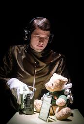 Vergrößerte Ansicht von Person mit Kopfhörern und weißen Handschuhen hält ein Funkgerät vor einem sitzenden Teddybär mit grünem Pullover auf einem Tisch