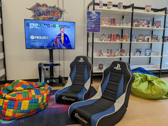 Vergrößerte Ansicht von Ein Foto vom Gaming-Bereich. Vor einem Fernseher mit Playstation 5- und Switch-Konsole stehen Gaming-Sessel und Sitzsäcke. Neben dem Fernseher ein Regal mit Videospielen.