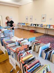 Vergrößerte Ansicht von Eine Frau blättert in einem Raum mit Tischen voller Bücher in einem Buch.