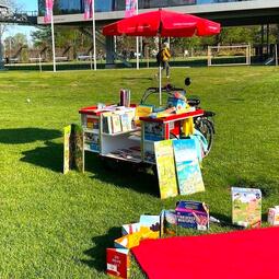 Vergrößerte Ansicht von Ein rotes Lastenrad mit Sonnenschirm und ausklappbarer Box mit Medien steht in einem sonnigen Park.