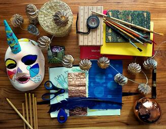 Vergrößerte Ansicht von Bastelmaterialien auf einem Tisch. Darunter eine Maske, Pinsel, Papier und Stifte.