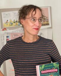 Vergrößerte Ansicht von Eine Frau mit Brille und Büchern in der Hand lächelt.