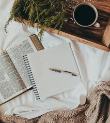 Vergrößerte Ansicht von ein Blick von oben auf eine Decke, auf der ein aufgeschlagenes Buch, ein Notizheft, ein Stift und ein Tablett liegen. Auf dem Tablett befinden sich eine gefüllte Kaffeetasse und ein Tannenzweig.
