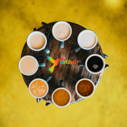 Vergrößerte Ansicht von Ein Blick von oben auf einen runden Tisch. Darauf stehen Tassen im Kreis, gefüllt mit unterschiedlich hellem und dunklem Kaffee. Der Hintergrund ist gelb. In der Mitte ein bunter Kolibri-Vogel neben dem Wort colibri. 