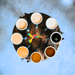 Vergrößerte Ansicht von Ein Blick von oben auf einen runden Tisch. Darauf stehen Tassen im Kreis, gefüllt mit unterschiedlich hellem und dunklem Kaffee. Der Hintergrund ist blau. In der Mitte befindet sich ein bunter Kolibri-Vogel neben dem Wort colibri.