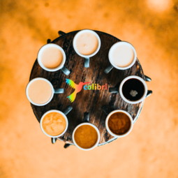 Vergrößerte Ansicht von Ein Blick von oben auf einen runden Tisch vor einem Hintergrund in orange. Darauf stehen Tassen im Kreis, gefüllt mit unterschiedlich hellem und dunklem Kaffee. In der Mitte ein bunter Kolibri-Vogel neben dem Wort colibri.