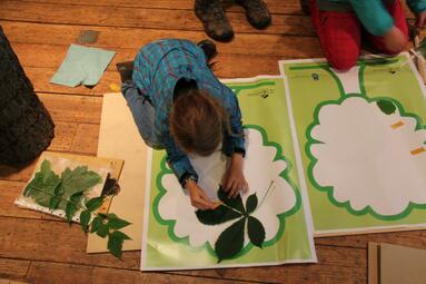 Vergrößerte Ansicht von Mädchen hat vor sich auf dem Boden ein grünes Plakat und ein Kastanienblatt liegen und bastelt
