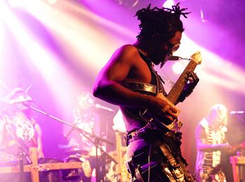 Während des PLANET EARS Festival 2023 tritt die Band Fulu Miziki in der Alten Feuerwache auf. Zu sehen ist ein in violettes Licht getauchter mit silbern glänzender Maske und kunstvollen Accessoires bekleideter Gitarrist auf der Bühne.