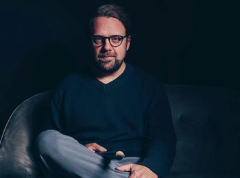 Ein Mann mit Brille sitzt auf einem Sofa. Der Raum ist dunkel, Schatten fällt auf sein Gesicht. Er hat die Beine überschlagen, in der Hand ein überdimensionierter Bleistift.