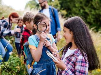 Klassenausflug im Grünen. Zwei Mädchen im Vordergrund. Die hintere hält eine große Lupe vors Gesicht. Beide lächeln.