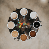 Ein runder, brauner Tisch. Darauf im Kreis Kaffeetassen, gefüllt mit unterschiedlich starkem Kaffee. In der Mitte ein bunter Colibri-Vogel mit dem Schriftzug "Colibri Online".