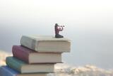 Auf einem Bücherstapel sitzende Miniaturfigur mit einem Fernglas in der Hand