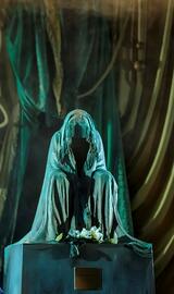 Eine Statue steht auf einem massiven Sockel, beleuchtet mit einem grünen Licht. Eine vorgebeugte Figur ist mit einem Hellen Tuch überdeckt.
