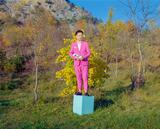 Konstantin Gropper steht auf einem hellblauen Würfel vor einer herbstlichen Baum- und Berglandschaft. Er trägt einen rosafarbenen Anzug und schaut erst, wärend er ein abstraktes, längliches Objekt hält.