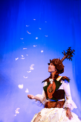 Eine Frau im herbstlich-winterlichen Theater-Kostüm sieht zu Federn hoch, die wie Schneeflocken rieseln.