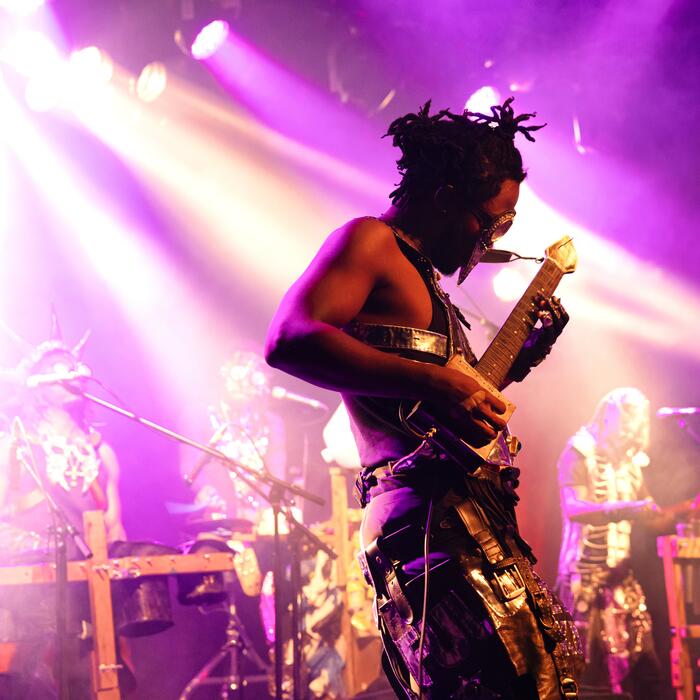 Während des PLANET EARS Festival 2023 tritt die Band Fulu Miziki in der Alten Feuerwache auf. Zu sehen ist ein in violettes Licht getauchter mit silbern glänzender Maske und kunstvollen Accessoires bekleideter Gitarrist auf der Bühne.