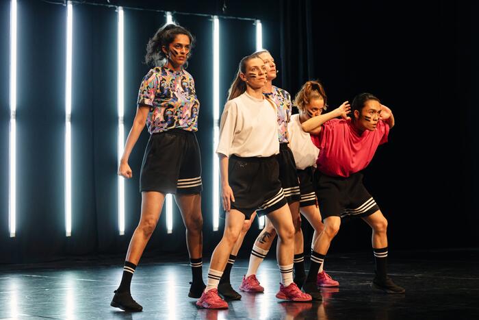 Während des Schwindelfrei Festival 2024 wird das Stück "Radical Cheerleading“ von Zufit Simon im EinTanzHaus präsentiert. Zu sehen sind fünf Personen, die in sportlicher Kleidung mit entschlossenem Blick auf der Bühne stehen.